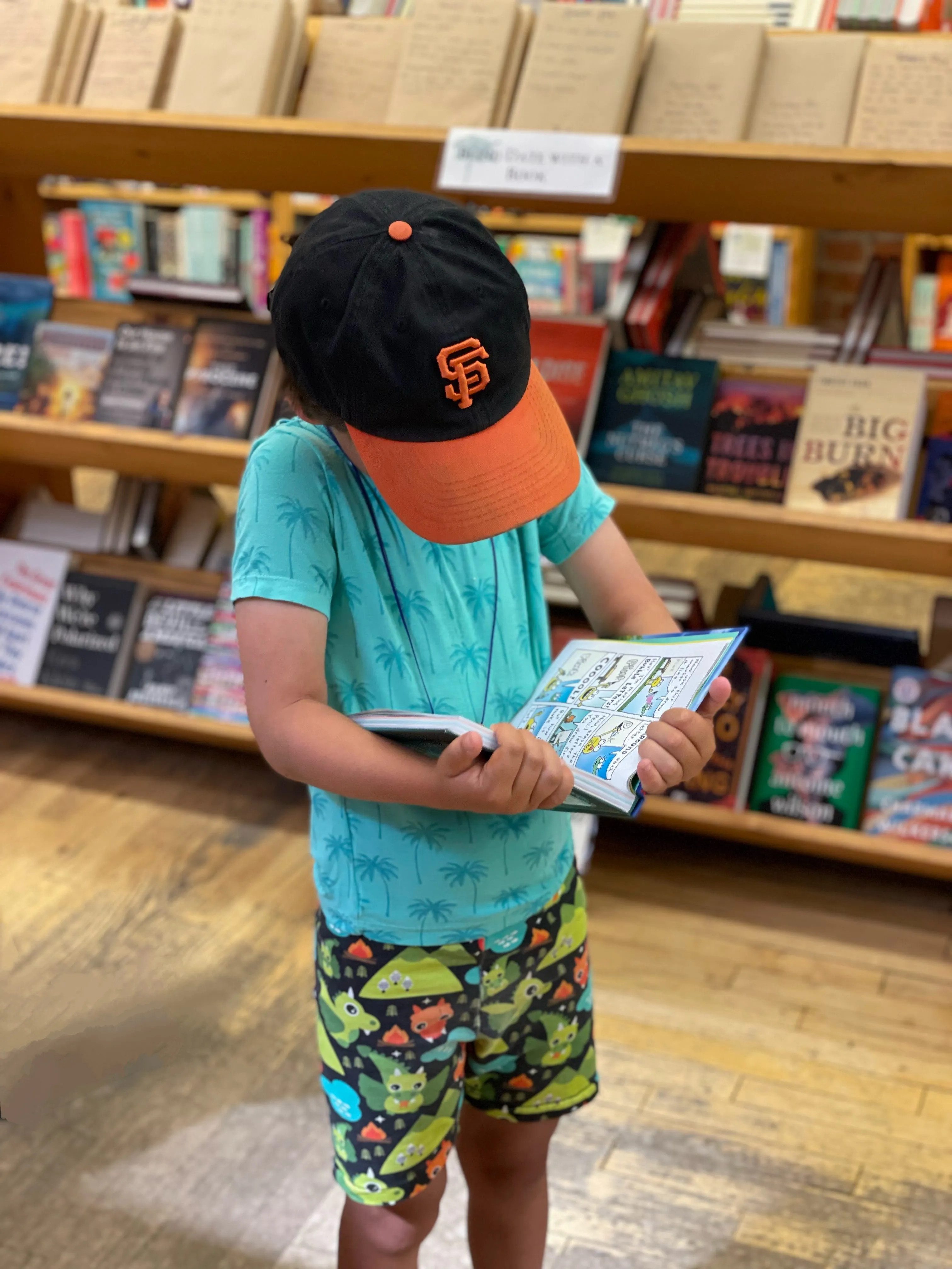 A small child wearing a hat that obscures their face is standing, reading a book. The shelf behind the child suggests they are in a library or bookstore.