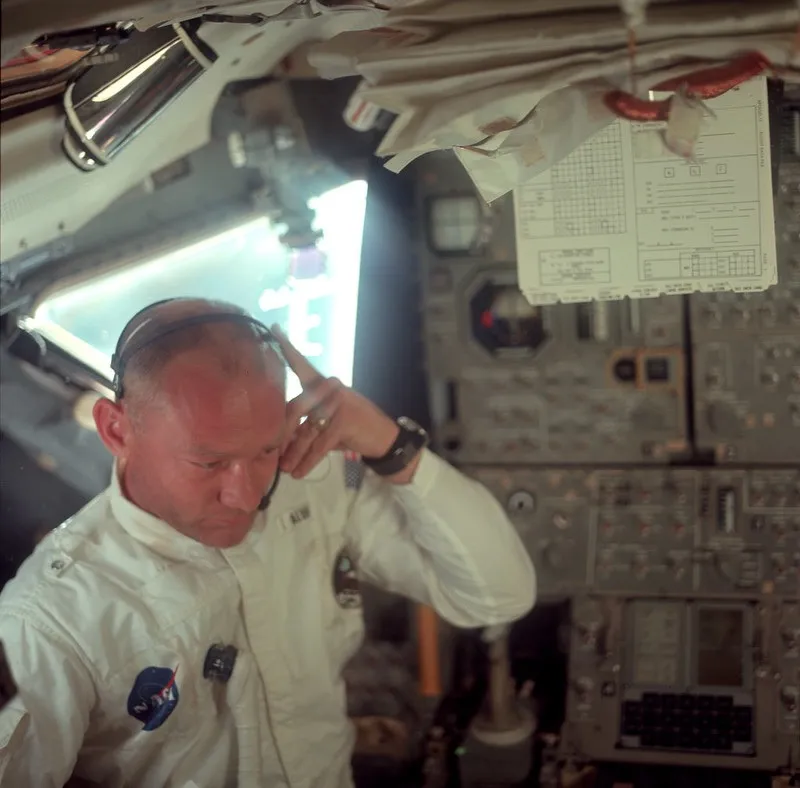 Buzz Aldrin, in the Apollo 11 capsule, with a bright window visible and many dials and switches behind him. He is wearing white clothing with NASA patches, but not a full space suit, and is focused on whatever is in front of him, out of frame.