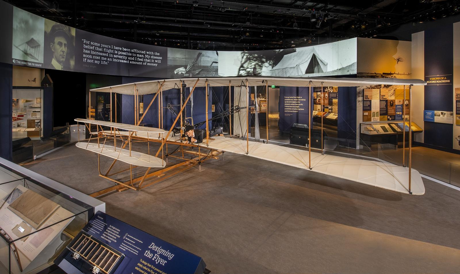 The 1903 Wright Flyer on display: a wood biplane with fabric-covered wings, a figure lying face down on the lower wing, and interpretive panels and historic photographs along the surrounding gallery walls